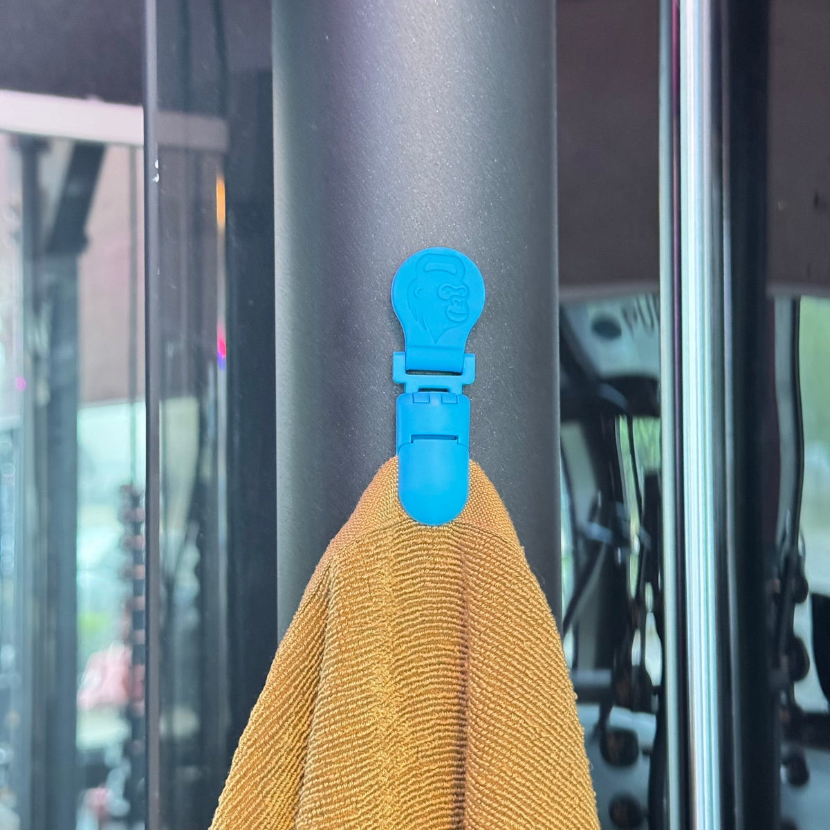 Nahaufnahme eines magnetischen Handtuchclips mit Affenlogo, der ein gelbes Gym Handtuch an einem Fitnessgerät hält